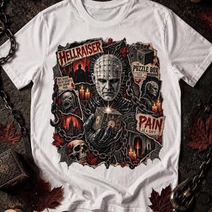 May include: A white t-shirt featuring a graphic print of the Hellraiser character Pinhead holding a puzzle box. The design includes text reading HELLRAISER, THE PUZZLE BOX, WE HAVE SUCH SIGHTS TO SHOW YOU, and PAIN IS A GIFT, surrounded by chains and skulls.