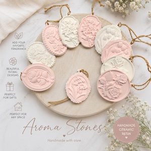May include: A collection of oval and round ceramic resin aroma stones featuring embossed floral designs in white and soft pink. Each piece includes a jute twine loop for hanging. The text reads: Aroma Stones, Handmade with care.