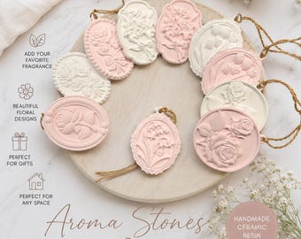 May include: A collection of oval and round ceramic resin aroma stones featuring embossed floral designs in white and soft pink. Each piece includes a jute twine loop for hanging. The text reads: Aroma Stones, Handmade with care.
