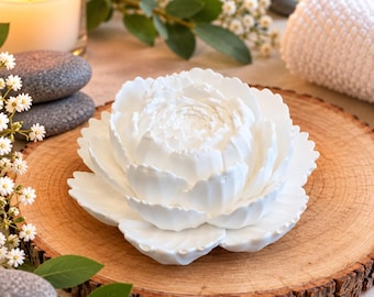 May include: A white ceramic flower sculpture with layered, rounded petals arranged in a blooming peony shape. The object features a smooth, matte finish and is centered on a circular wooden slice with visible bark edges and grain.
