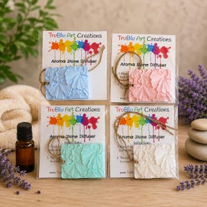 May include: Four square aroma stone diffusers by TruBlu Art Creations in light blue, pink, teal, and white. Each stone features a raised ginkgo leaf pattern, a jute hanging string, and is packaged with instructions on a card with a colorful paint splatter logo.