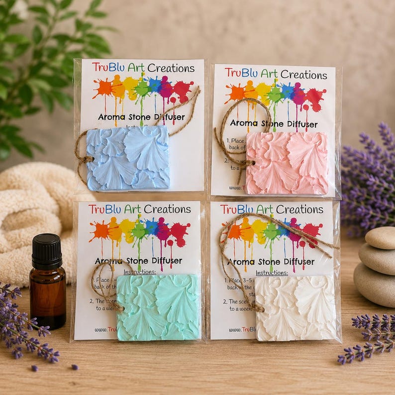 May include: Four square aroma stone diffusers by TruBlu Art Creations in light blue, pink, teal, and white. Each stone features a raised ginkgo leaf pattern, a jute hanging string, and is packaged with instructions on a card with a colorful paint splatter logo.