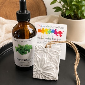 May include: A rectangular white aroma stone diffuser with a raised daisy floral pattern, featuring a jute twine hanger. Beside it sits a 4 oz amber glass bottle of Brooklyn Botany peppermint essential oil with a black dropper cap.