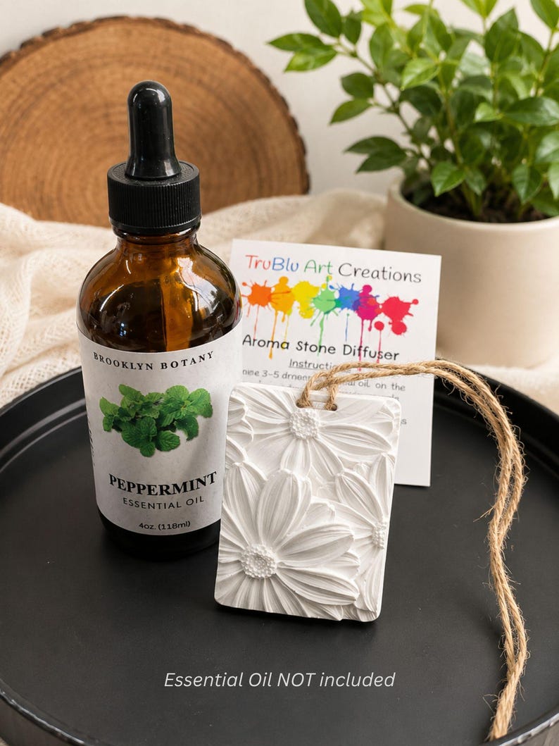 May include: A rectangular white aroma stone diffuser with a raised daisy floral pattern, featuring a jute twine hanger. Beside it sits a 4 oz amber glass bottle of Brooklyn Botany peppermint essential oil with a black dropper cap.