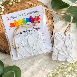 May include: Two rectangular white aroma stone diffusers featuring a raised ginkgo leaf texture. One diffuser is inside a clear plastic bag with a TruBlu Art Creations label, while the other sits beside it. Both have attached brown twine hanging loops.