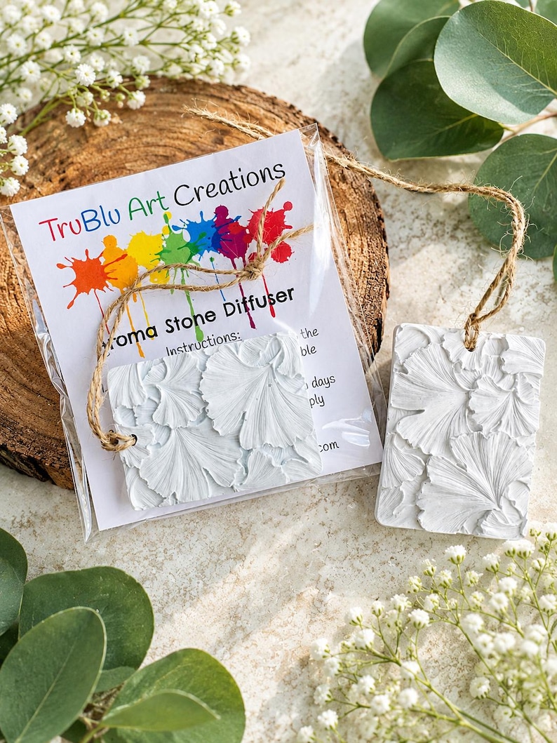 May include: Two rectangular white aroma stone diffusers featuring a raised ginkgo leaf texture. One diffuser is inside a clear plastic bag with a TruBlu Art Creations label, while the other sits beside it. Both have attached brown twine hanging loops.