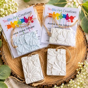 May include: Four rectangular aroma stone diffusers with embossed floral patterns. Two are packaged in clear plastic with cards labeled TruBlu Art Creations Aroma Stone Diffuser, while two are loose. All feature attached jute twine hanging loops.