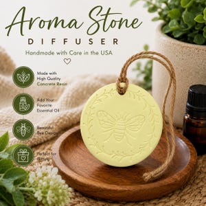 May include: A round, pale yellow concrete resin aroma stone diffuser featuring an embossed bee design surrounded by a leafy wreath. The diffuser hangs from a brown jute twine cord and is displayed on a small, circular wooden tray.