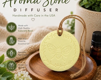 May include: A round, pale yellow concrete resin aroma stone diffuser featuring an embossed bee design surrounded by a leafy wreath. The stone hangs from a brown jute twine cord and is displayed on a small, circular wooden tray.