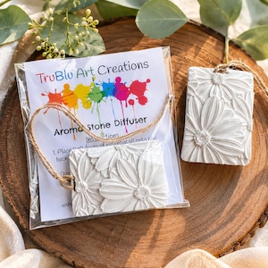 May include: Two rectangular white aroma stone diffusers featuring a raised daisy floral pattern. One diffuser is inside a clear plastic bag with a TruBlu Art Creations instruction card, while the other sits beside it, both attached to brown twine.