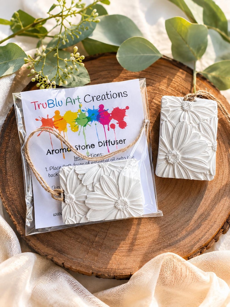 May include: Two rectangular white aroma stone diffusers featuring a raised daisy floral pattern. One diffuser is inside a clear plastic bag with a TruBlu Art Creations instruction card, while the other sits beside it, both attached to brown twine.
