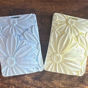 May include: Two rectangular plaques featuring a raised, three-dimensional floral pattern of daisies. One plaque is light blue and the other is pale yellow. Each plaque has a small rectangular cutout at the top center for hanging purposes.