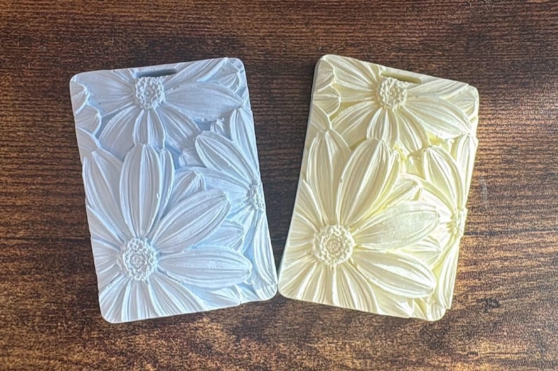 May include: Two rectangular plaques featuring a raised, three-dimensional floral pattern of daisies. One plaque is light blue and the other is pale yellow. Each plaque has a small rectangular cutout at the top center for hanging purposes.