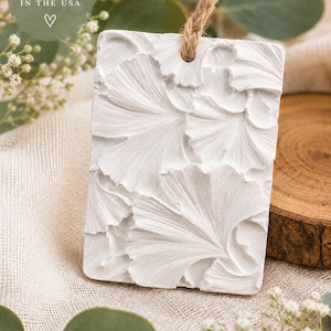 May include: A rectangular white hanging ornament featuring a raised, textured ginkgo leaf pattern. The piece includes a hole at the top threaded with a natural jute twine cord. Text on the image reads: HANDMADE WITH CARE IN THE USA.