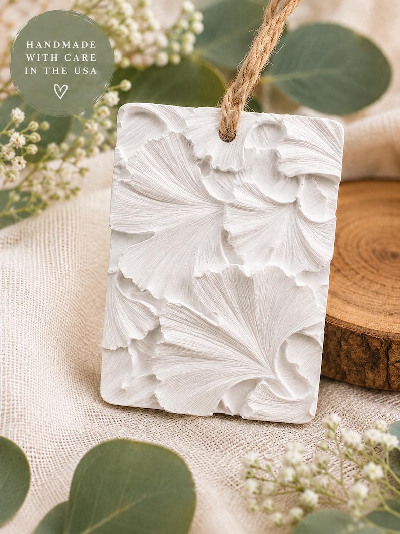 May include: A rectangular white hanging ornament featuring a raised, textured ginkgo leaf pattern. The piece includes a hole at the top threaded with a natural jute twine cord. Text on the image reads: HANDMADE WITH CARE IN THE USA.