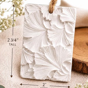 May include: A rectangular white ornament featuring a raised ginkgo leaf pattern on the front. The piece includes a brown jute twine loop at the top for hanging. It measures 2 3/4 inches tall and 2 inches wide as indicated by the text.
