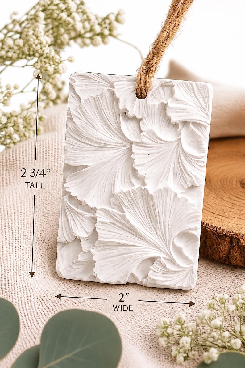 May include: A rectangular white ornament featuring a raised ginkgo leaf pattern on the front. The piece includes a brown jute twine loop at the top for hanging. It measures 2 3/4 inches tall and 2 inches wide as indicated by the text.