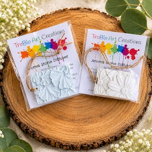May include: Two rectangular aroma stone diffusers in clear plastic packaging with twine hangers. One stone is light blue and the other is white, both featuring embossed floral patterns. The labels read TruBlu Art Creations Aroma Stone Diffuser.