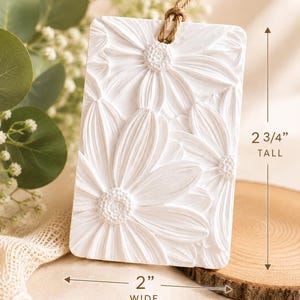 May include: A rectangular white plaque with a raised relief floral pattern of daisies. The plaque features a brown twine loop at the top for hanging. Dimensions are clearly marked as 2 3/4 inches tall and 2 inches wide on the image.