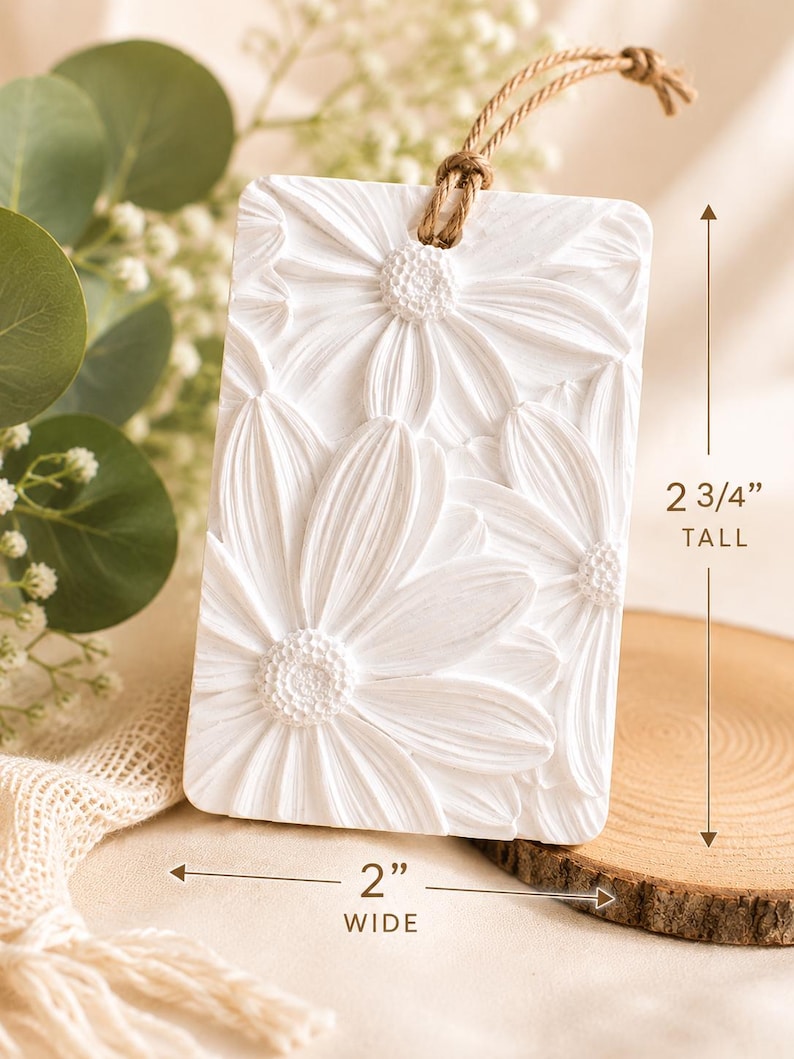 May include: A rectangular white plaque with a raised relief floral pattern of daisies. The plaque features a brown twine loop at the top for hanging. Dimensions are clearly marked as 2 3/4 inches tall and 2 inches wide on the image.