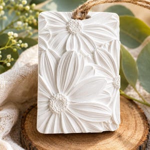May include: A rectangular white ceramic hanging ornament featuring a detailed relief of multiple daisy flowers with textured centers and smooth petals. A natural jute twine loop is threaded through a hole at the top of the white floral plaque.