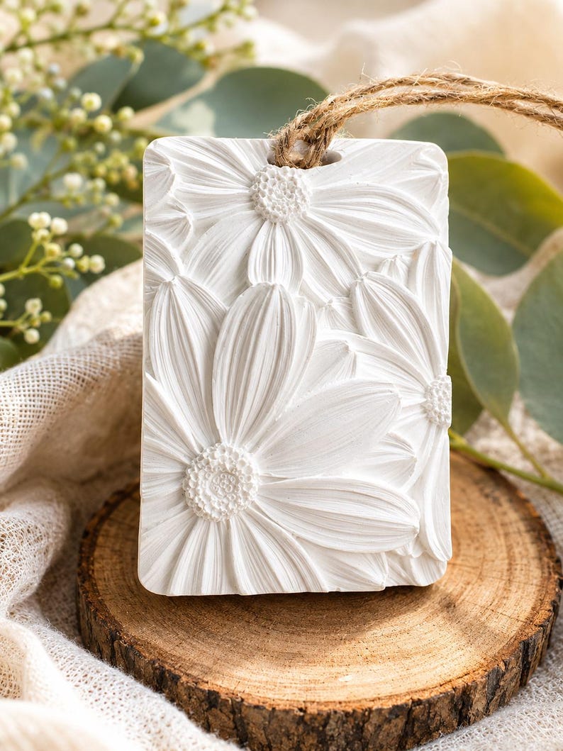 May include: A rectangular white wax melt or hanging ornament featuring a detailed, raised relief of multiple daisy flowers. The piece has a smooth, matte finish and includes a small hole at the top threaded with a natural brown jute twine.