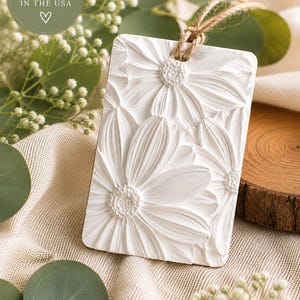 May include: A rectangular white hanging ornament featuring a raised, textured floral relief design of daisies. The ornament includes a natural jute twine loop at the top for hanging. A circular green sticker in the corner reads HANDMADE WITH CARE IN THE USA.