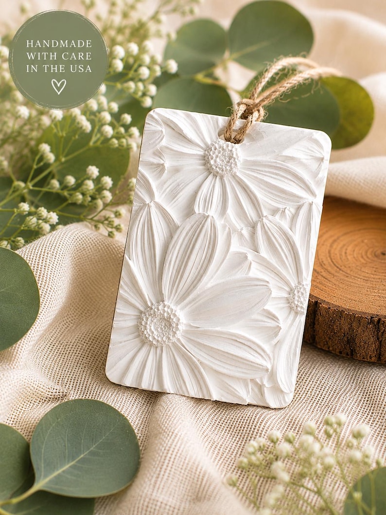 May include: A rectangular white hanging ornament featuring a raised, textured floral relief design of daisies. The ornament includes a natural jute twine loop at the top for hanging. A circular green sticker in the corner reads HANDMADE WITH CARE IN THE USA.