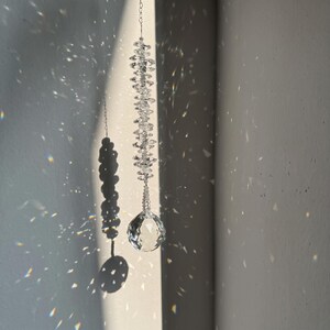 Pode incluir: Um apanhador de sol de cristal transparente pendurado numa fina corrente prateada. O ornamento apresenta uma pilha vertical de pequenas contas de cristal facetado encimada por um grande prisma de cristal esférico e multifacetado.