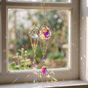 Könnte beinhalten: Ein hängender Sonnenfänger mit einem kugelförmigen, facettierten Kristall in einem goldfarbenen Metalldrahtkäfig, der an einer Goldkette hängt. Ein kleinerer, schillernder Kristalltropfen hängt darunter und wirft bunte Lichtreflexe auf die Fläche.