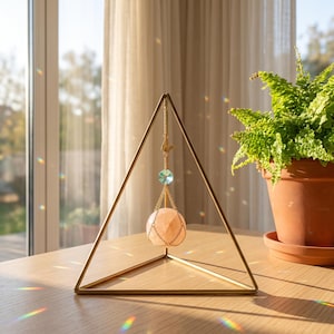 Pode incluir: Uma estrutura triangular de metal dourado que sustenta um ornamento suspenso. O ornamento apresenta uma pedra esférica rosa-pálida envolta numa rede de correntes douradas, com uma pequena conta azul-clara facetada e um pendente de pássaro.