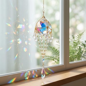 Pode incluir: Um apanhador de sol de cristal suspenso com uma grande esfera iridescente facetada suspensa por correntes douradas. O design inclui gotas e contas de cristal transparente menores, criando refrações de luz e prismas coloridos.