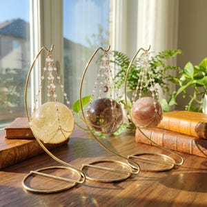 Può includere: Tre cristalli sferici nelle tonalità giallo, marrone scuro e bianco rosato, ciascuno sospeso in un supporto in metallo dorato. Le sfere sono sostenute da delicate catenelle di perline con piccoli ciondoli a forma di mezzaluna.