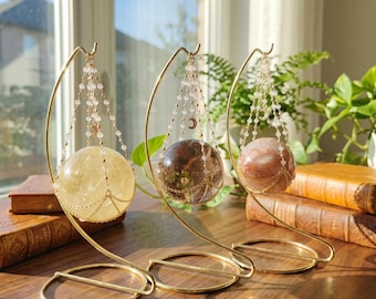 May include: Three spherical crystals in yellow, dark brown, and pinkish-white hues, each suspended in a gold-toned metal stand. The spheres are held by delicate beaded chains featuring small crescent moon charms, creating a hanging display.