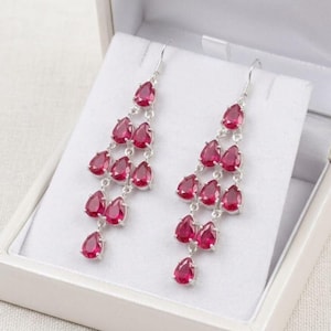 Puede incluir: Un par de pendientes de araña de plata con múltiples piedras rojas en forma de lágrima dispuestas en un diseño en cascada. Cada pendiente cuelga de un gancho de plata y muestra una serie de gemas rojas facetadas unidas entre sí.