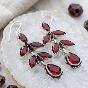 May include: A pair of silver dangle earrings featuring multiple faceted deep red garnet gemstones. Each earring is composed of several marquise-cut stones arranged in a leaf-like pattern, terminating in a single pear-shaped drop with a twisted metal border.