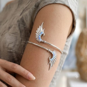 May include: A silver-toned metal arm cuff featuring two symmetrical wing-shaped ends. Each wing is embellished with a series of small, round, iridescent opalescent stones that reflect light with blue and white hues along the curved metal structure.