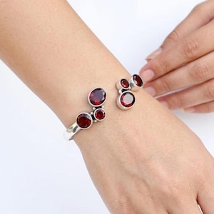 May include: A silver cuff bracelet featuring five round, deep red garnet gemstones of varying sizes. The stones are set in polished silver bezels, with three stones clustered on one end and two stones on the other, creating an open-ended design.