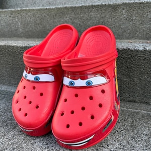 Può includere: Un paio di zoccoli rossi vivaci con un design grafico ispirato al personaggio Saetta McQueen del film Cars. Le scarpe presentano occhi bianchi e blu stampati sulla parte anteriore, fori di ventilazione e un cinturino posteriore sagomato.