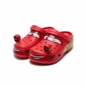 Pode incluir: Um par de tamancos vermelhos sem atacadores com um design de personagem inspirado num carro de corrida. Os sapatos incluem gráficos de olhos azuis e brancos no topo, uma boca sorridente no para-choques frontal e o número 95 em amarelo.