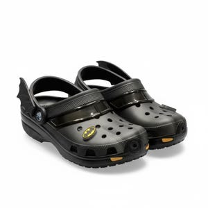 Pode incluir: Um par de tamancos pretos com tema Batman. Os sapatos possuem cabedal perfurado, tira de calcanhar giratória com detalhes de asas de morcego, um emblema do Batman amarelo e preto e pequenos detalhes retangulares amarelos na sola.