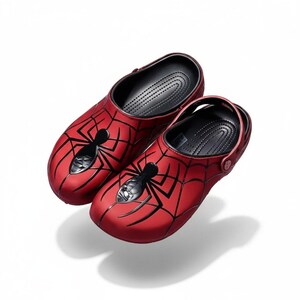 May include: A pair of red clogs featuring a black spider web pattern across the top and a prominent black spider emblem centered on the toe area. The interior footbed is black with a textured surface, and each shoe includes a pivoting heel strap.