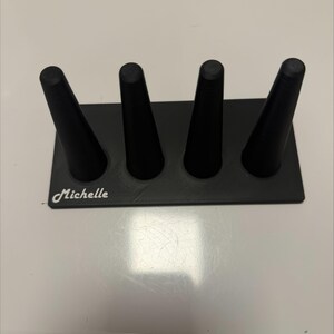 May include: A black ring holder stand featuring a rectangular base with four upright conical posts. The name Michelle is printed in white script lettering on the bottom left corner of the base. The object has a matte finish and a structured design.