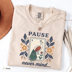 May include: An ivory crew neck t-shirt featuring a graphic of a playing card with a peacock illustration, the number one, and the text PAUSE never mind. The design is flanked by dark blue leaves and orange flowers, with a Comfort Colors brand label.