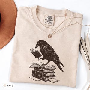 May include: A beige short-sleeve t-shirt featuring a dark brown graphic of a raven standing on a stack of three books while holding an open book in its claw. The inner neck label reads Comfort Colors, Heavyweight Garment Dye, 100% Cotton, Made in Honduras, S-P-CH.
