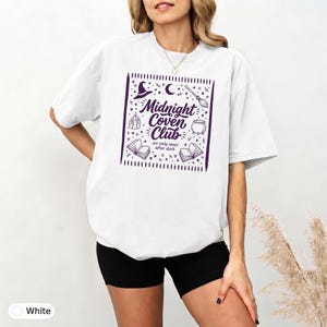 Pode incluir: Uma t-shirt branca de manga curta com uma impressão gráfica roxa. O design inclui o texto "Midnight Coven Club" e "we only meet after dark," rodeado por ilustrações de um chapéu de bruxa, lua crescente, vassoura, velas, caldeirão e livros.