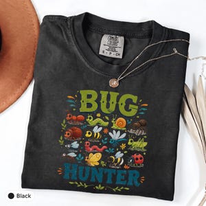 May include: A black t-shirt featuring a graphic print of various colorful insects and the text "BUG HUNTER" in green and blue. The shirt has a Comfort Colors brand label at the neck and is paired with a delicate gold-toned chain necklace.