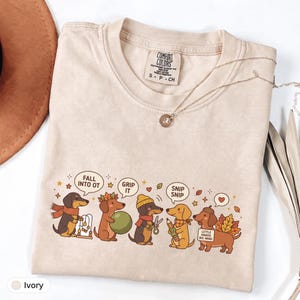 May include: An ivory crewneck t-shirt featuring a graphic of five dachshunds engaged in occupational therapy activities. The dogs are shown with speech bubbles containing the text FALL INTO OT, GRIP IT, and SNIP SNIP, along with a sign reading LITTLE HANDS BIG WINS.