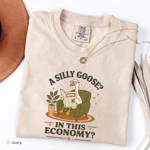 May include: An ivory t-shirt featuring a graphic of a white goose wearing glasses, sitting in a green armchair, reading a newspaper titled HONK. The text reads A SILLY GOOSE? IN THIS ECONOMY? with a potted plant and star accents in green and orange.
