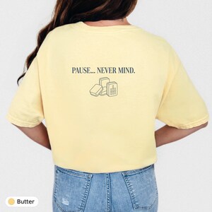May include: A butter yellow short-sleeve t-shirt featuring the text PAUSE... NEVER MIND. printed in dark blue on the back. Below the text is a simple dark blue line art illustration of three small rectangular boxes or cases arranged together.